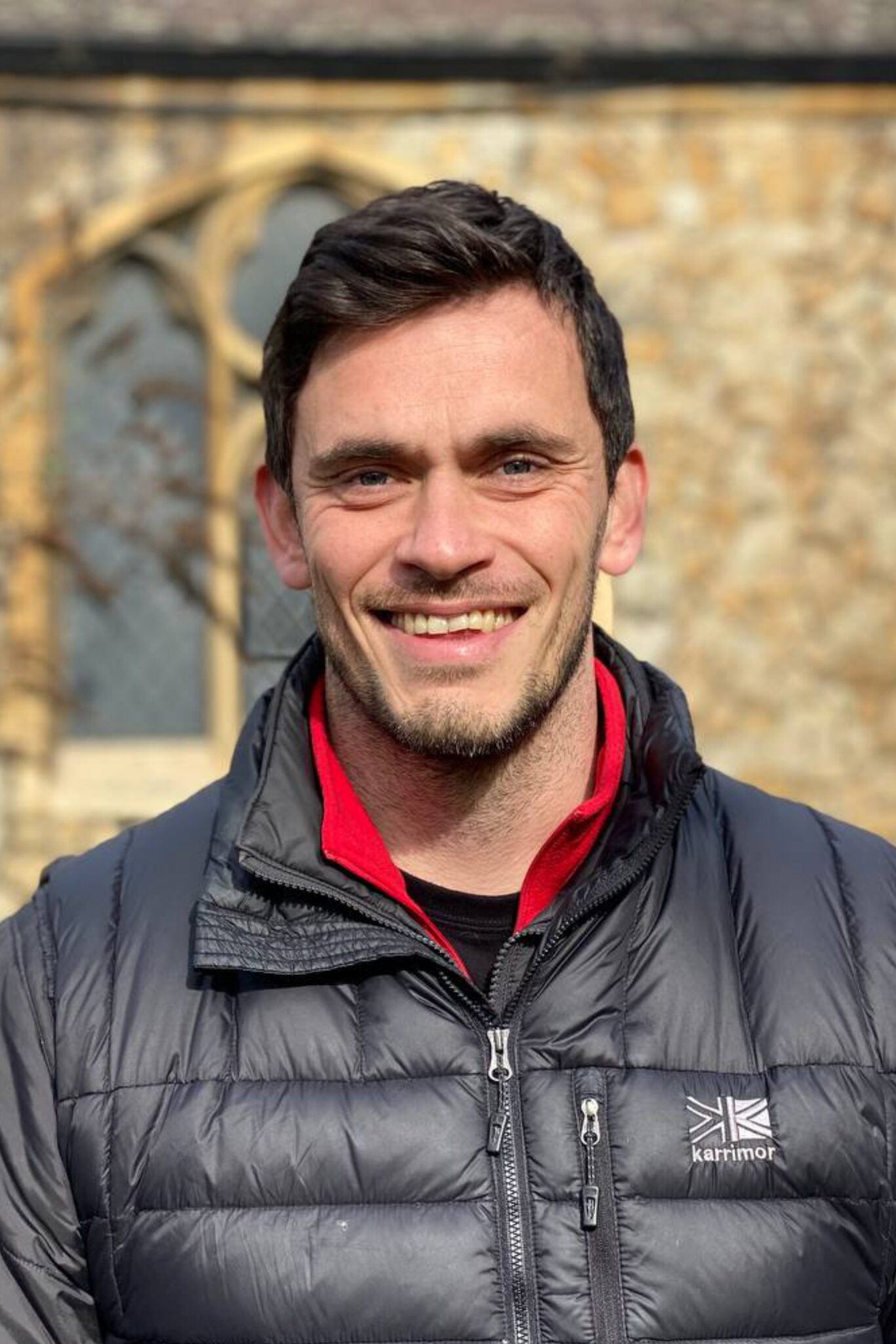 white male with dark hair wearing outdoor jacket infront of church wall